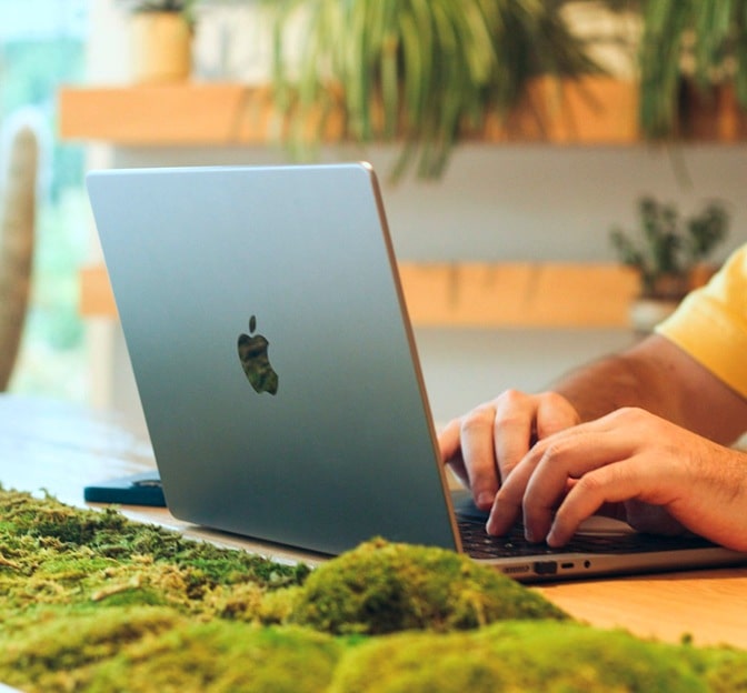 A person typing on a MacBook, with a lush green moss surface and potted plants in the background, creating a natural work environment.