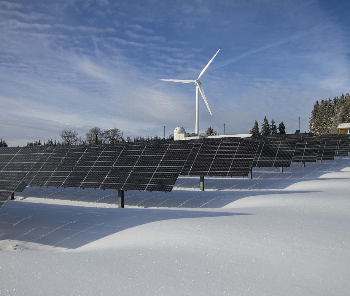 A snowy landscape featuring solar panels in the foreground and a wind turbine in the background, under a clear blue sky.